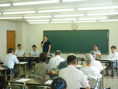 黒板のある部屋に机が並べられており、正面に立って話をしている男性と椅子に座って話を聞いている参加者たちの写真