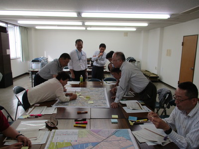 机に置かれた大きい地図に屈みこみながらペンで書き込んでいる女性と、周囲から様子を見ている関係者たちの写真