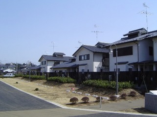 生垣とフェンスに囲まれた奥に壁が白色で平屋建てと2階建ての建物が繋がっている清水台住宅の写真
