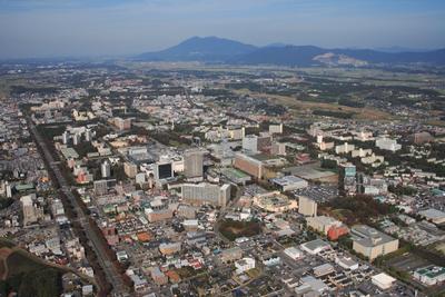青い空を背景に、奥に高い山と田畑、全体をビルやマンション、建物が広がる筑波研究学園都市の様子を、上空から撮影した写真