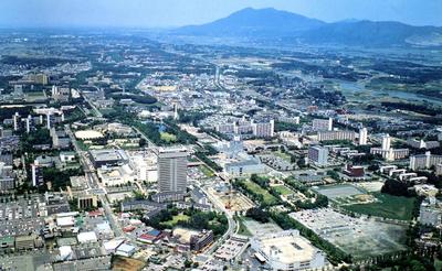 青い空を背景に、奥に高い山、全体を緑の樹木とビルやマンション、建物が広がる筑波研究学園都市の様子を、上空から撮影した写真