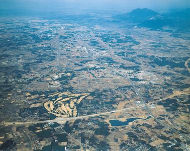 奥に高い山、全体を低い山と緑と黄土色から成る田畑が広がり、それらを横断する長い道路がある筑波研究学園都市の様子を、上空から撮影した写真