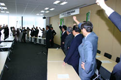 10人以上のスーツを着た年配の男性が、カタカナのハの字に並べられた長机の前で、左手を腰に当て右手拳を上に掲げている様子を撮影した写真
