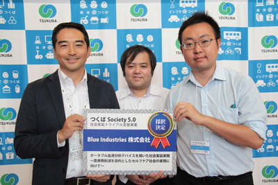 Blue Industries株式会社の2名の男性のうちの1人と市長が一緒に採択決定と書かれたボードを持っている表彰の模様の写真