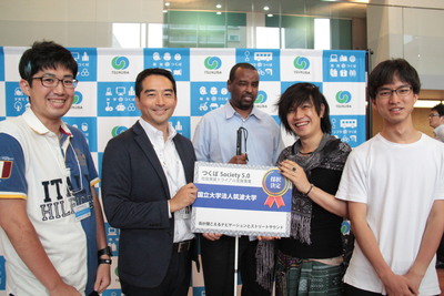 筑波大学の学生4名のうちの1人と市長が一緒に採択決定と書かれたボードを持っている表彰の模様の写真