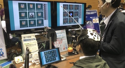 椅子に座っている頭上に脳波の機械をつけた男性と横に立って機械を操作している男性が2台の画面を見ている実証実験の様子の写真