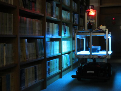 暗い部屋の中、ライトを転倒させながら本棚の間を巡回しているUV-C紫外線照射 自動巡回ロボットの写真