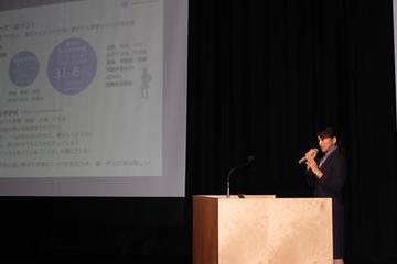 舞台上の暗幕に映し出されたプレゼン資料と演壇で話をしている筑波大学の学生の写真