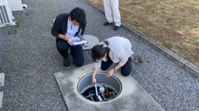 マンホールの蓋を開けて中の機械を調べている調査員と横でメモを取っている調査員の写真