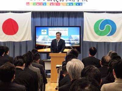 萩生田文部科学大臣が前でスピーチをしている写真。大臣の左側には日本の国旗が、右側にはつくば市の市章が掲げられている。