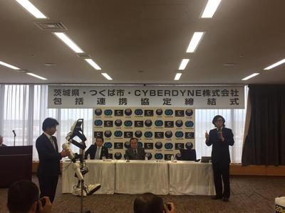 「茨城県・つくば市・CYBERDYNE株式会社包括連携協定締結式」と書かれたパネルの前で男性が話しているのを聞いている参加者の写真