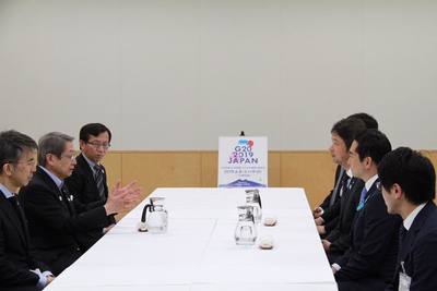 黒いスーツの男性達が白いテーブルを左右で囲むように椅子に腰かけ話している様子の写真