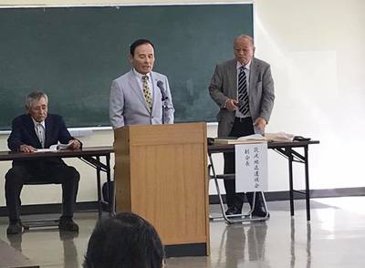 筑波地区遺族会総会で登壇して話す薄いグレーのスーツを着た男性と関係者が映っている写真