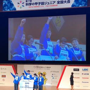ステージの上で青いユニフォームを着た子供5人が写っている写真。ステージの後ろの壁には「第7かい科学の甲子園ジュニア全国大会」と書かれている。