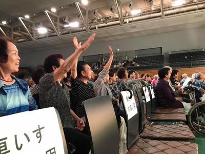 椅子が並べられた大きく広い会場で、座りながら手をあげる大人数の人達を撮影した写真。