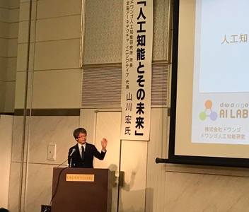 「「人工知能とその未来」山川 宏 氏」と書かれた懸垂幕の横で講演を行う山川氏の写真