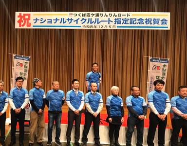 「つくば霞ヶ浦りんりんロード 祝 ナショナルサイクルルート指定記念祝賀会」と書かれたステージで、青い服を身につけて整列している人達の写真。