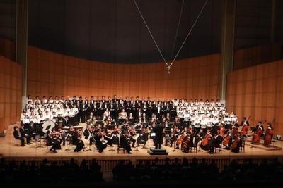 写真：つくばで第九（全体画像）舞台前方にオーケストラ、後方に合唱隊が並んでいる。中央に黒いスーツを着た男性、左右に白い服を着た女性