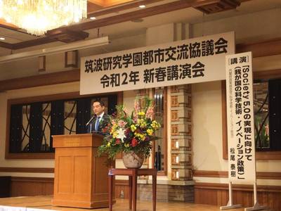 「筑波研究学園都市交流協議会 令和2年 新春講演会」と書かれた看板が掲げられたステージでスピーチをしている男性の写真。