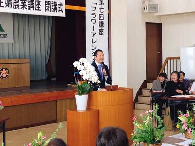 教壇でマイクをもって話をしている男性の写真。教団には白い大きな花も置かれている。