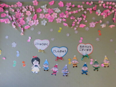 壁面にハートの形の枠に「ごにゅうがく、ごしんきゅうおめでとうございます」と書かれた文字と折り紙で作成した桜の花や白雪姫と七人の小人が飾られている写真