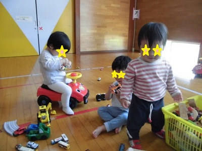 3名の小さな子どもが赤色の車のおもちゃに乗ったり、ミニカーを触ったりして遊んでいる写真