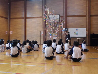 体育館で座っている多くの子どもたちの前で先生がお話をしている様子。先生の横には七夕の笹飾りがある。