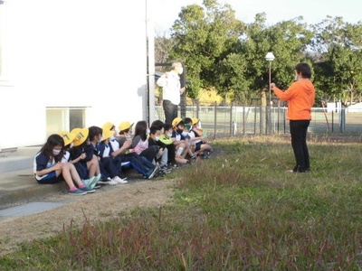建物の壁の近くに並んで座り、先生の話を聞いている子供たちの写真