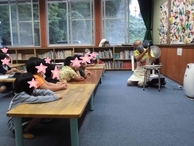 床に腰を下ろしている沢山の子供たちの前で読み聞かせをしている先生の写真
