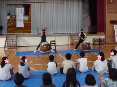 体育館で和太鼓をバチで叩いている2人の男性と演奏を見ている先生と子供たちの写真