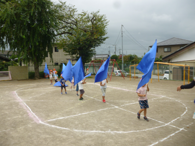 園庭を青い大きな旗を持って一列に歩く園児の写真