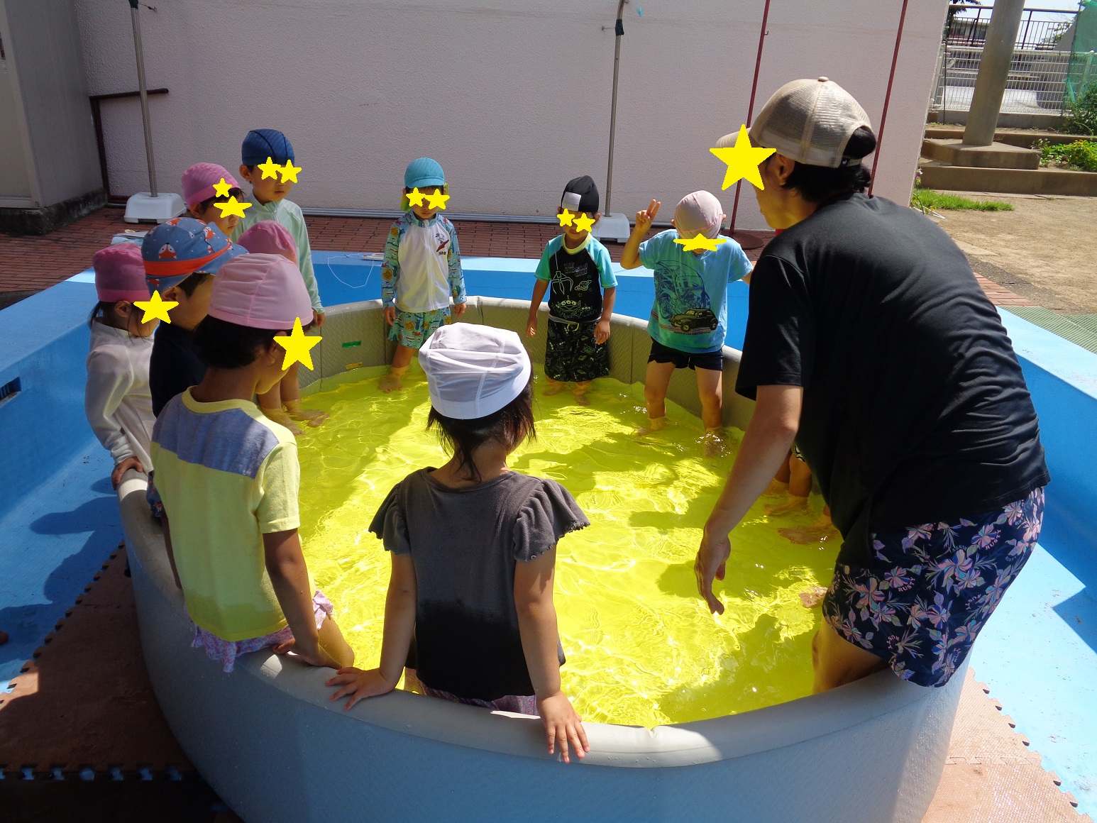 楕円形のプールに子どものくるぶしほどの水をため、水着やtシャツを着た子どもと大人たちが立っている