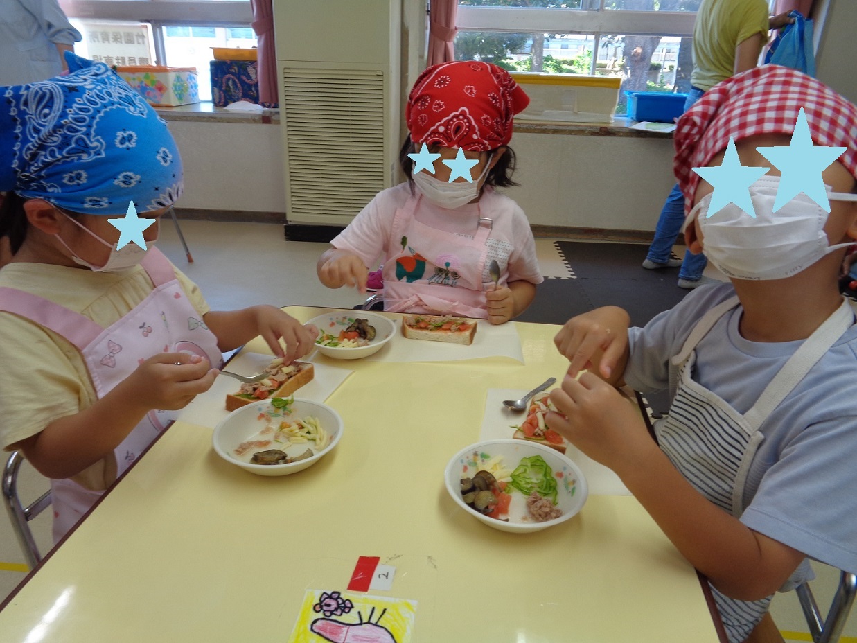 テーブルの周りに座り三角巾とエプロンを身に着けた子どもたちが、パンに切った野菜をのせている。