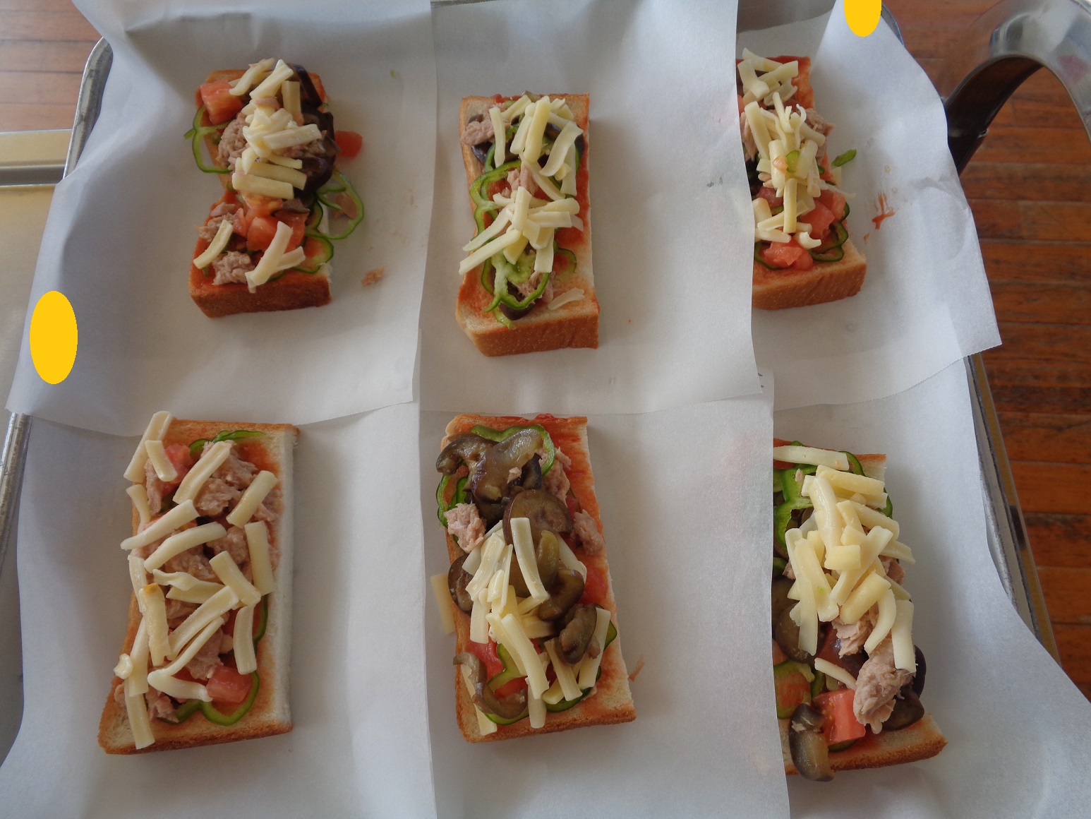 野菜とチーズをのせピザパンにするために食パン半分の大きさのパンが6枚並べて紙の上に置いてある