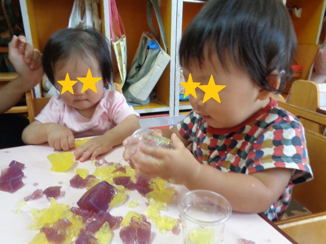 子どもたちが座ってテーブルの上にある黄色と紫色の寒天ゼリーをつまんだりカップを持っている