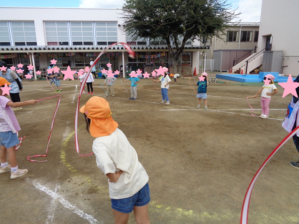 オレンジの帽子の子どもがリボンを持って踊るのをピンクの帽子を被った子どもたちが真似して踊っている。