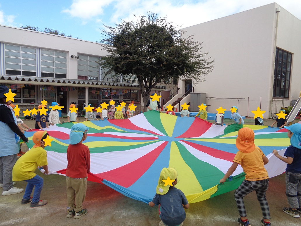 青い帽子とオレンジの帽子を被っている子どもたちが大きな布を広げて遊んでいる。