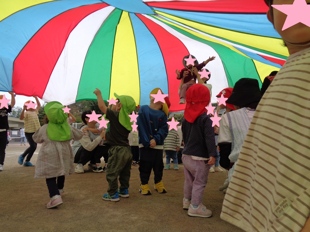 黄緑色の帽子と赤色の帽子を被った子どもたちが大きな布の下で遊んでいる。