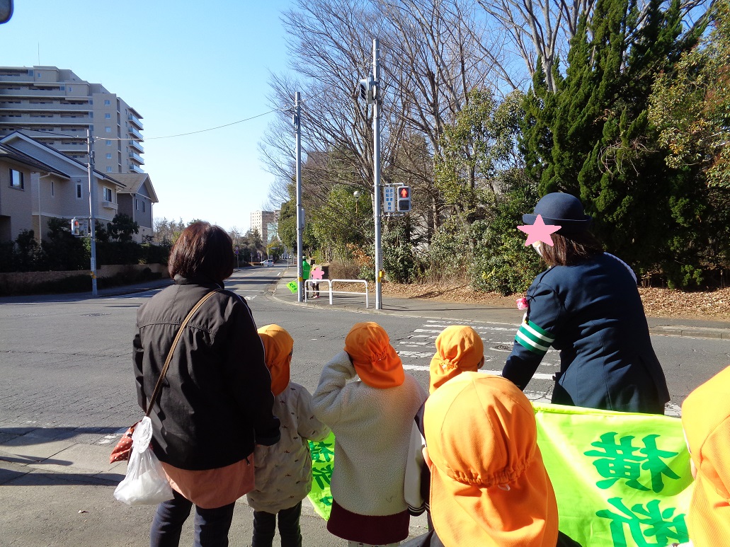 オレンジ色の帽子を被った子どもたちと大人が横断歩道の前で赤信号を見ている