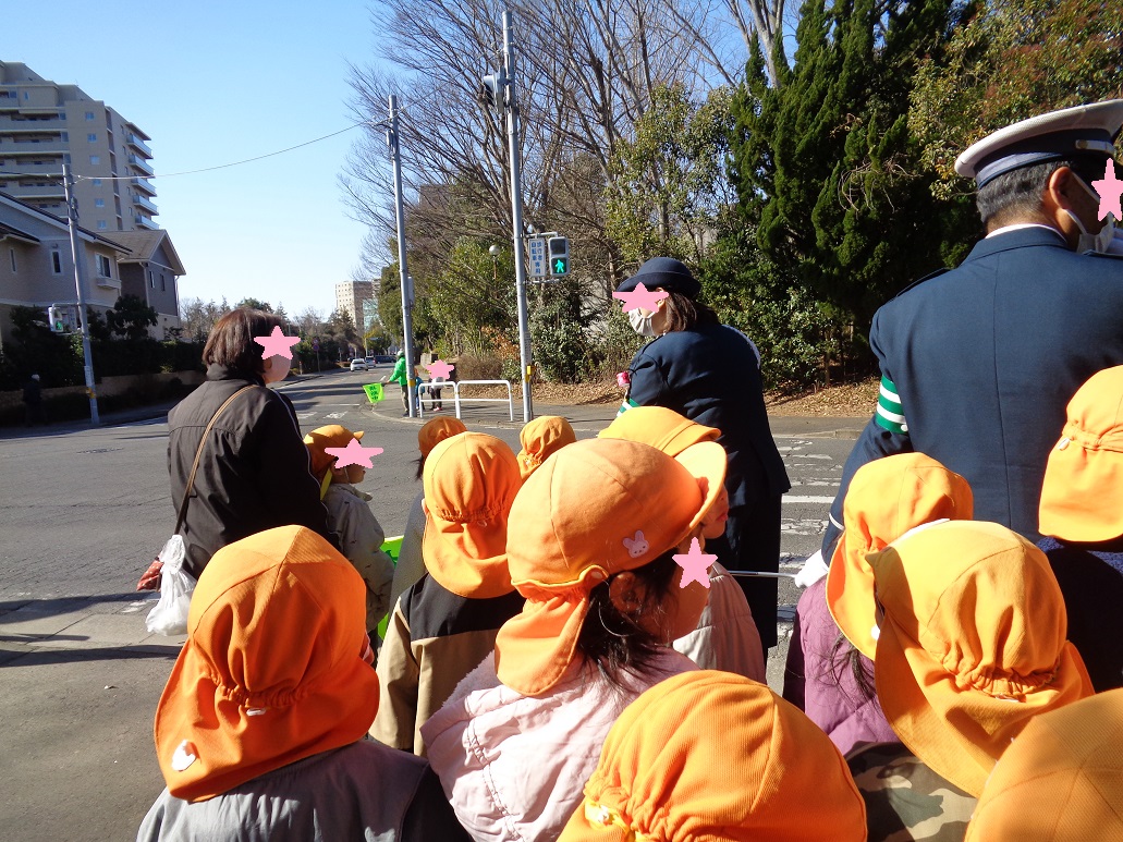 オレンジ色の帽子を被った子どもたちと大人が横断歩道の前で青信号を見ている