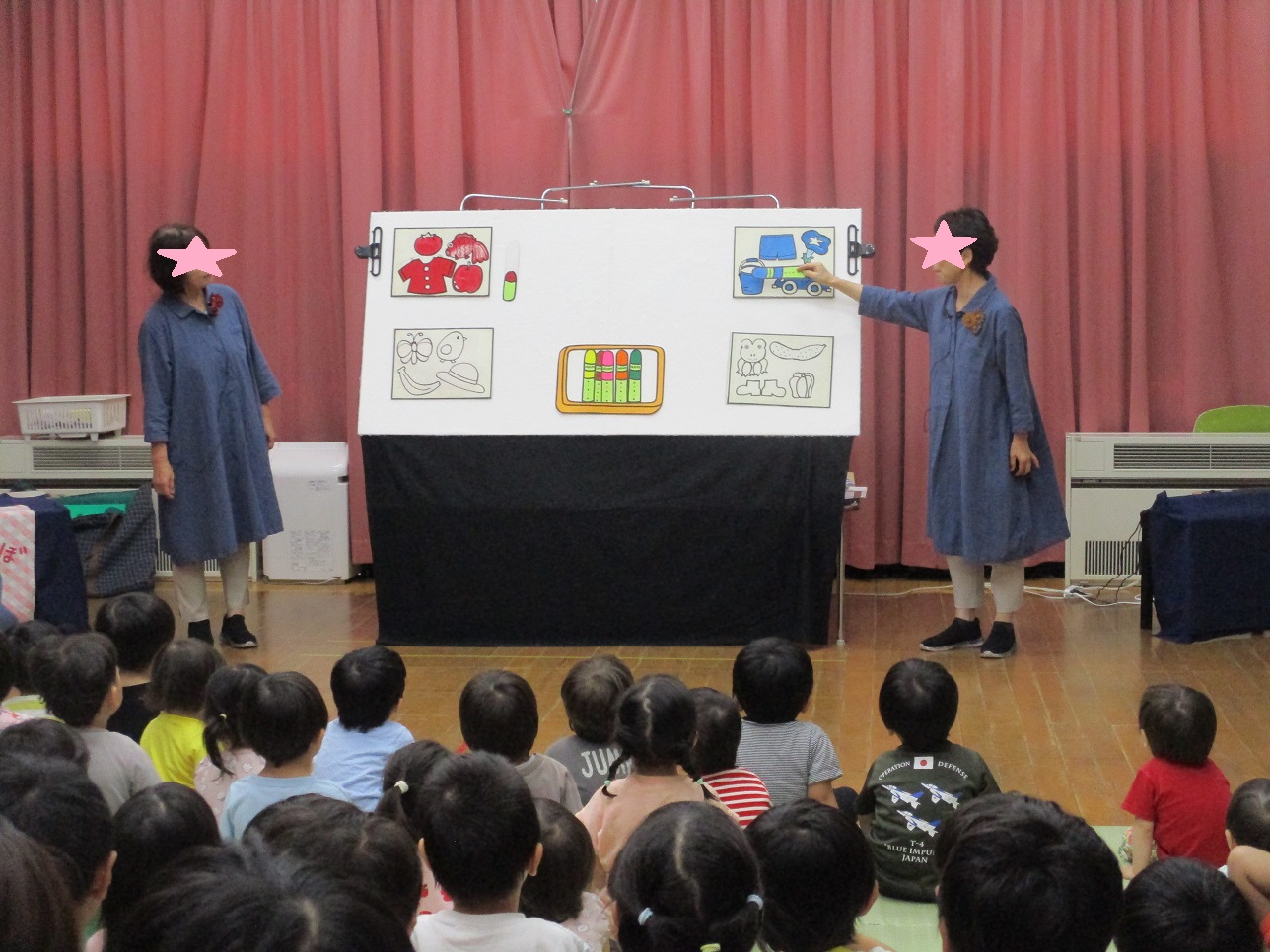 白い板にイラストをはり両脇に大人が話、それを見ている子供たち
